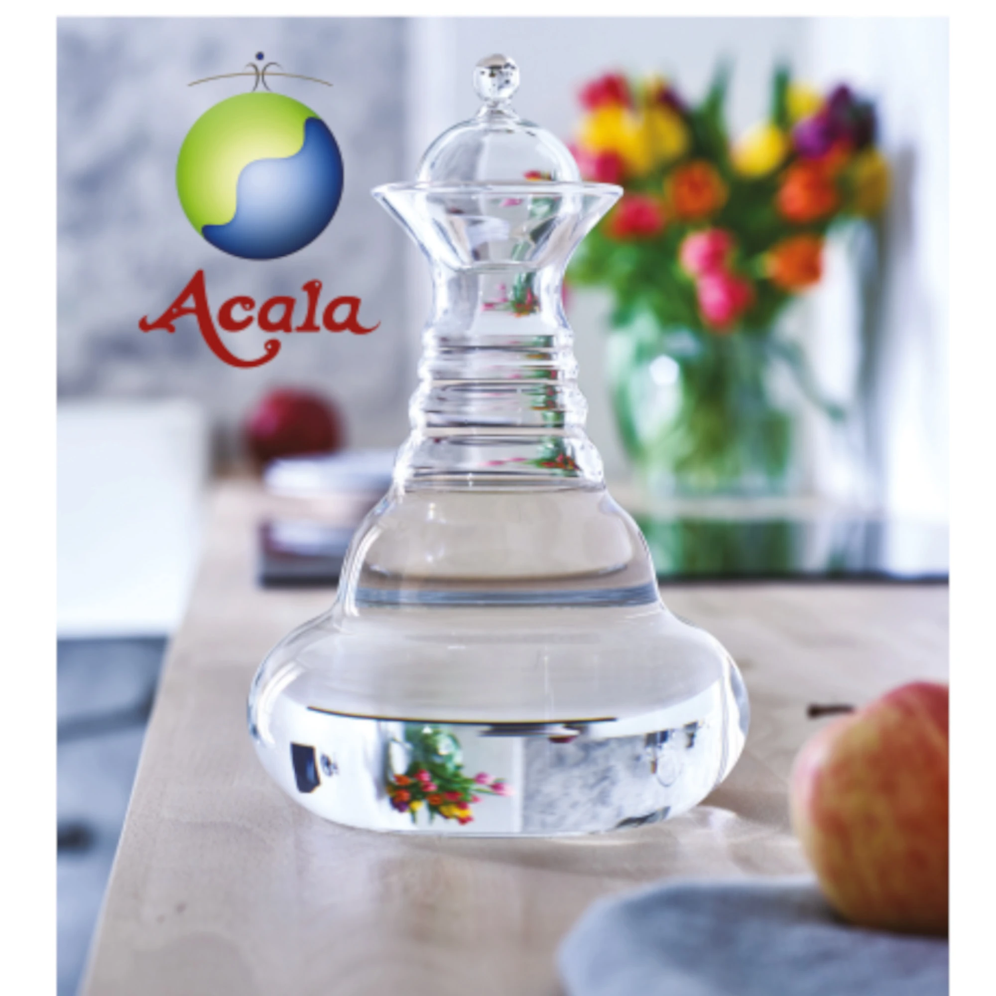 Zu sehen ist die Alladin Glas Karaffe von Acala mit Glasdeckel. Sie steht auf einem Tisch, man sieht Obst und einen bunten Blumenstrauß. Links neben der Karaffe sieht man das Acala Logo.