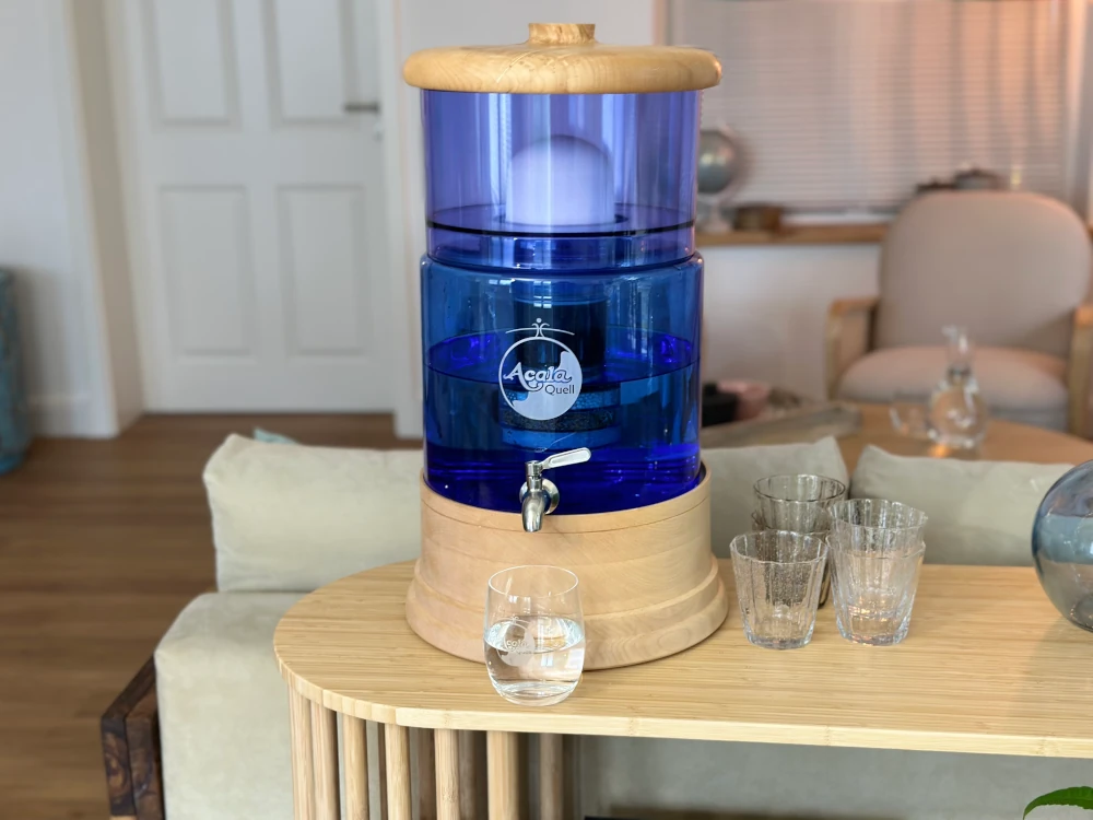 Acala Standwasserfilter Smart mit blauem Glas und Deckel und Sockel aus Ahorn Echtholz steht auf einem kleinen Tisch, Wasser läuft aus dem Edelstahl Wasserhahn in ein darunter stehendes Trinkglas. Im Hintergrund ein Wohnzimmer. 
