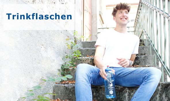 Zu sehen ist ein junger Mann der auf einer Steintreppe sitzt und lächelt. In der Hand hält er eine Tritan Trinkflasche mit einem Sportdeckel. Er ist Mitte zwanzig, hat kurze braune Haare, trägt ein weißes Shirt und eine blaue Jeans.