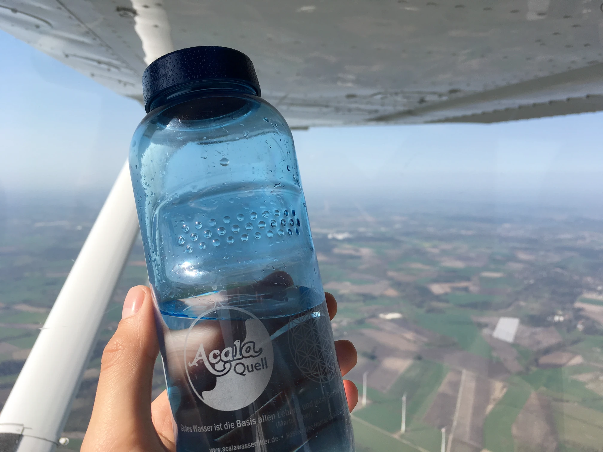 Tritanflasche in einer Hand die vor einem Fenster im Flugzeug ist und man sieht die Landaschaft aus dem Flugzeug