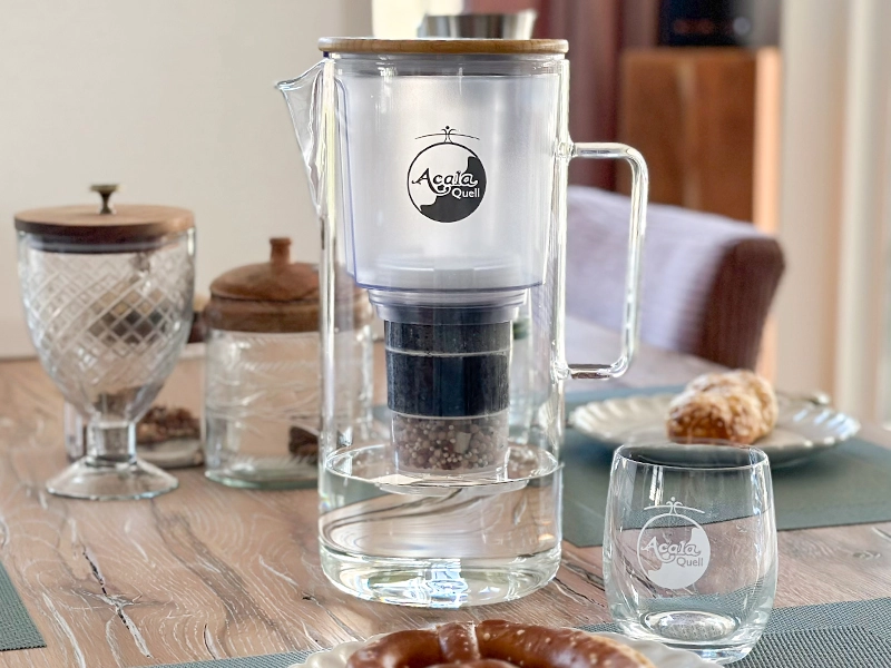 Zu sehen ist die Acala Glas Kanne Silizia auf einem braunen Holztisch. Es ist Wasser in der Kanne, daneben steht ein Glas mit Acala Logo und man sieht weitere Glasschalen mit Holzdeckel und Teller mit Brötchen darauf.