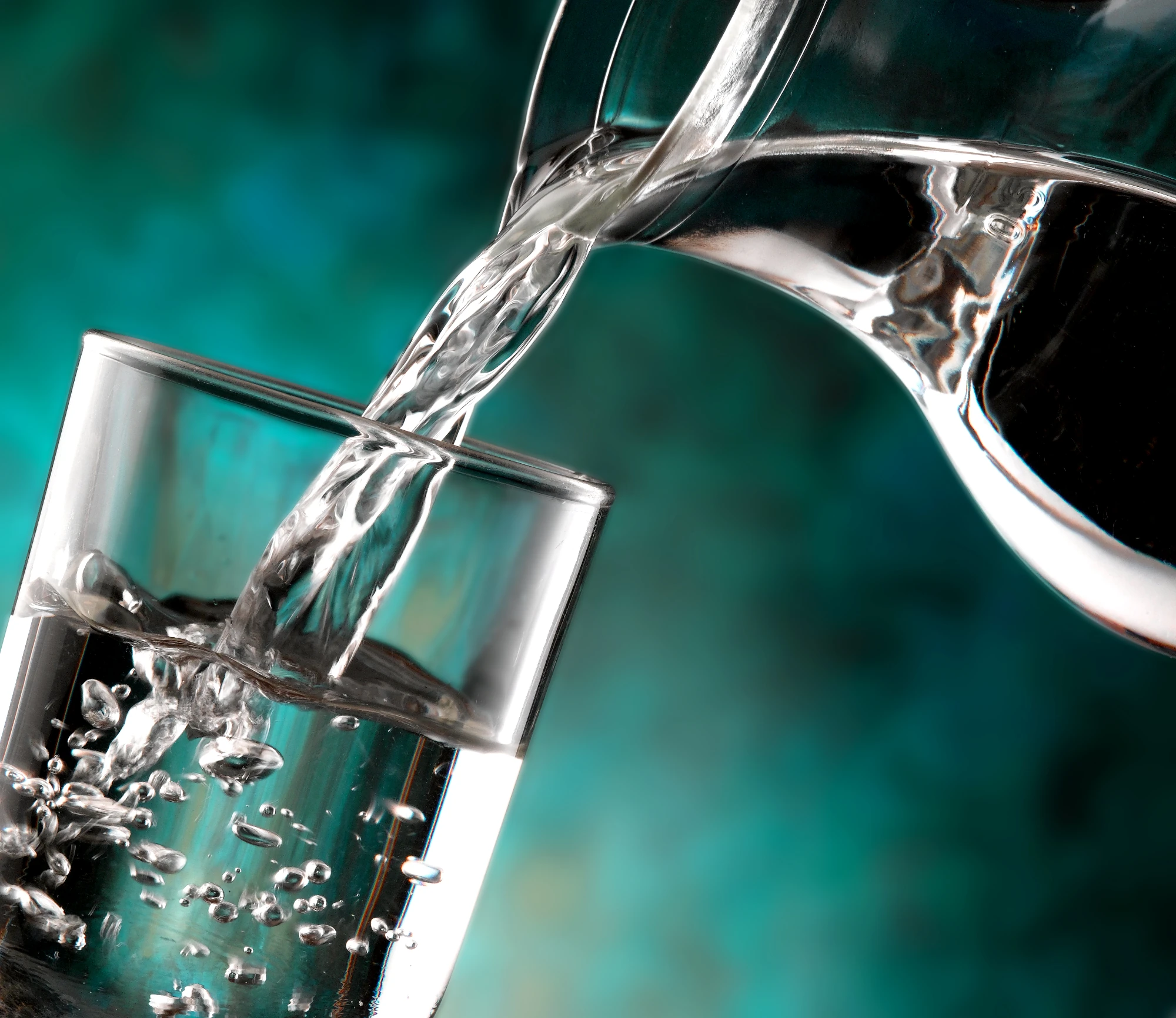 Klares Wasser wird aus einer Karaffe in ein Glas gegossen, vor einem dunkelgrünen Hintergrund