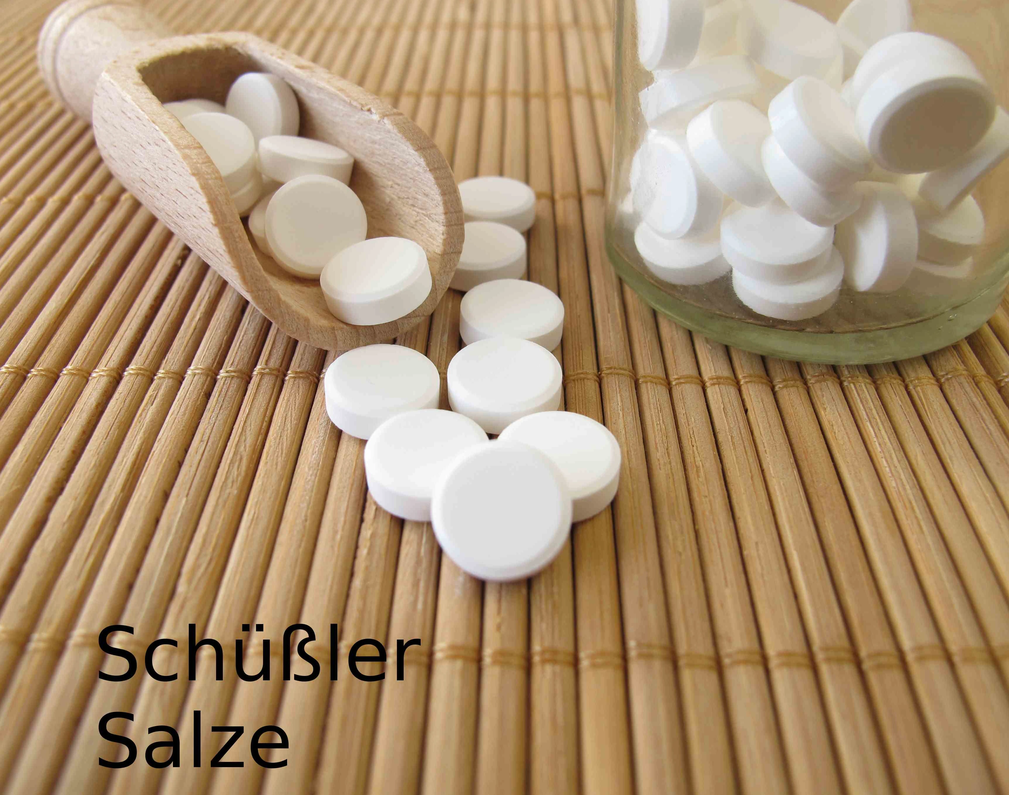 Schüsslersalze in Form von weißen Pillen auf einem Holzlöffen und in einem klaren Glas auf einer Bambusmatte